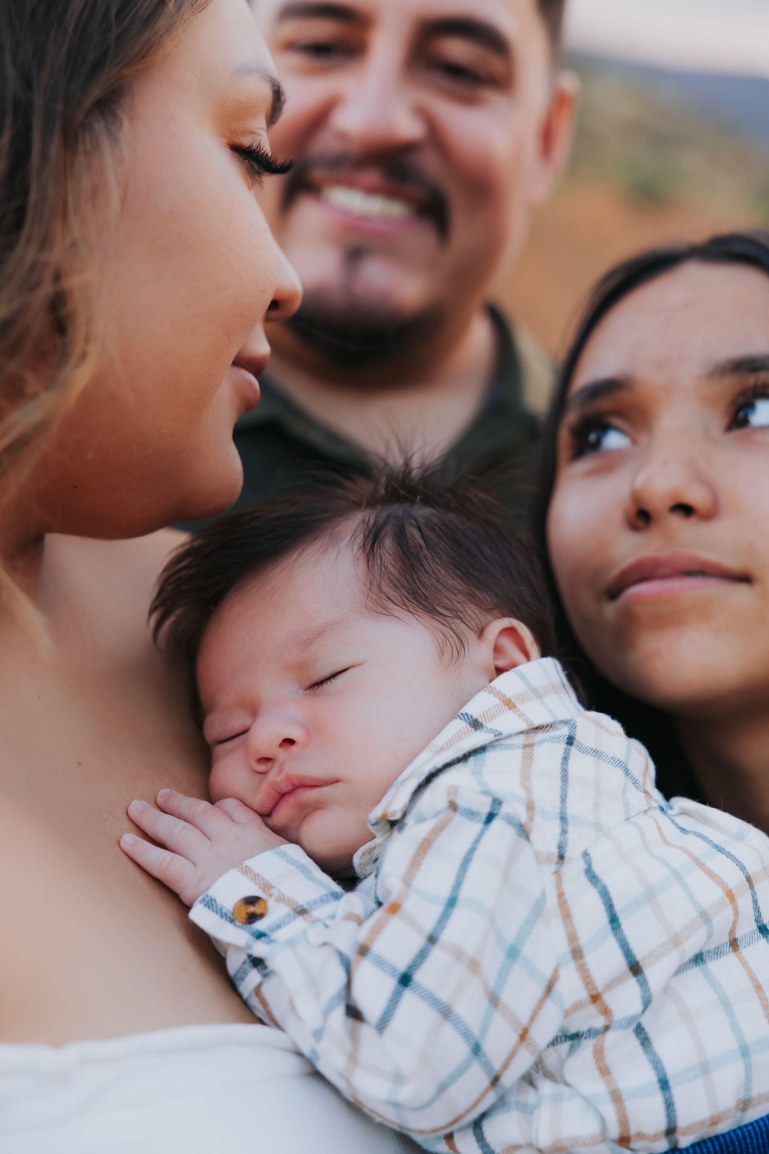 A sleeping infant wearing a checkered shirt is held lovingly in an intimate family portrait outdoors.