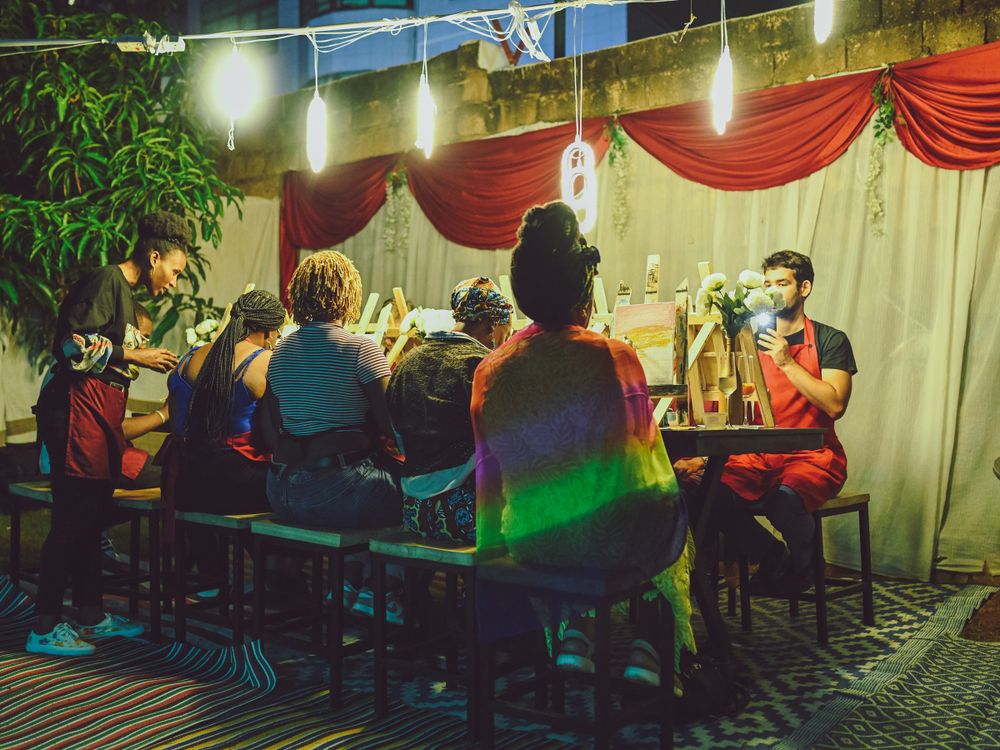 Group of people painting at an evening art class with string lights overhead.