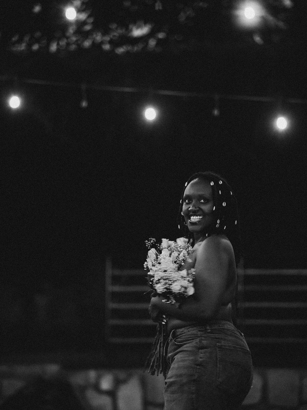 A figure holding a bouquet poses under string lights at night.