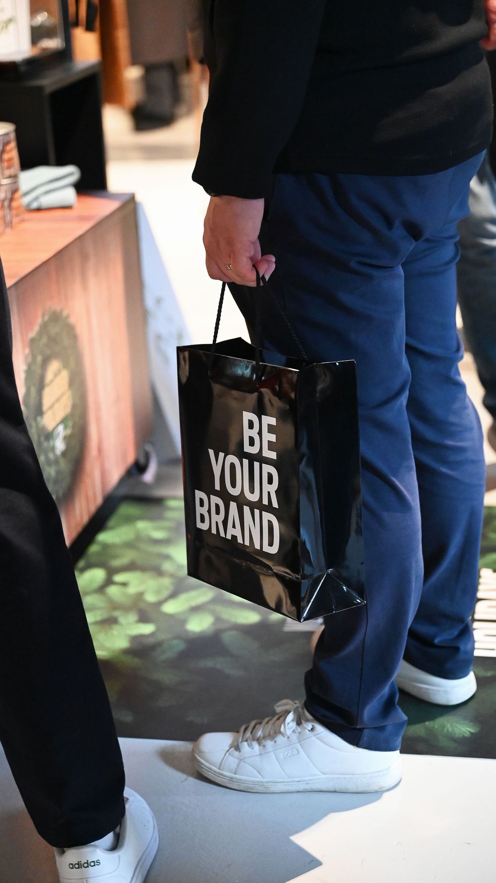 Black shopping bag with 'BE YOUR BRAND' text carried alongside white sneakers and blue jeans.