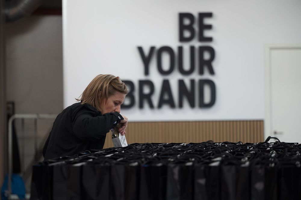 A person works at a desk with black bottles while a wall sign reads 'BE YOUR BRAND' in the background.