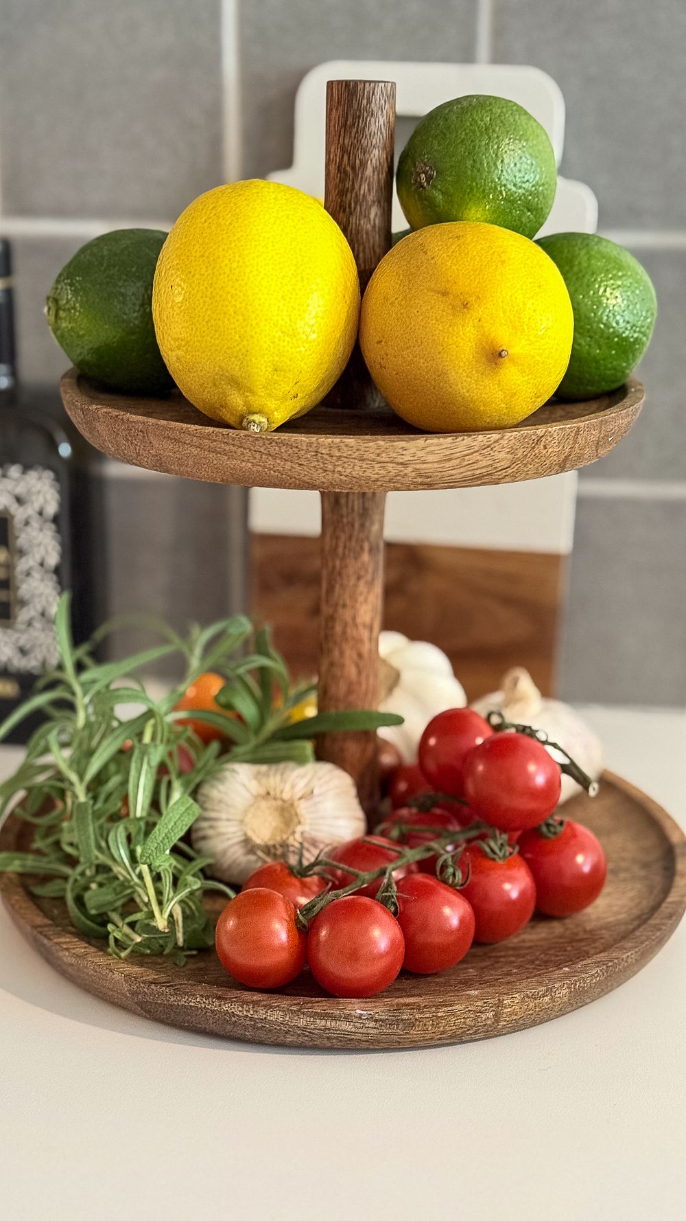 A wooden tiered serving stand displays fresh lemons, limes, cherry tomatoes, garlic, and herbs on a white counter.