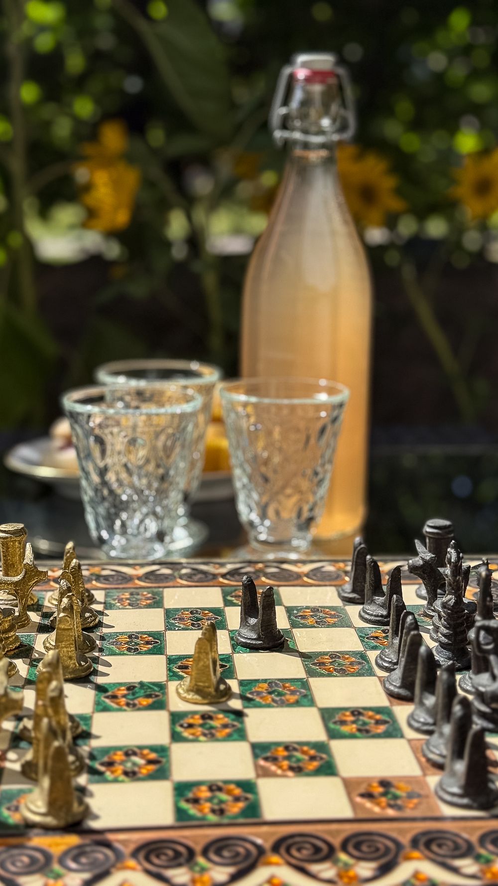 A vintage chess board with ornate pieces and floral pattern design is arranged with bottles and glasses in soft focus background.