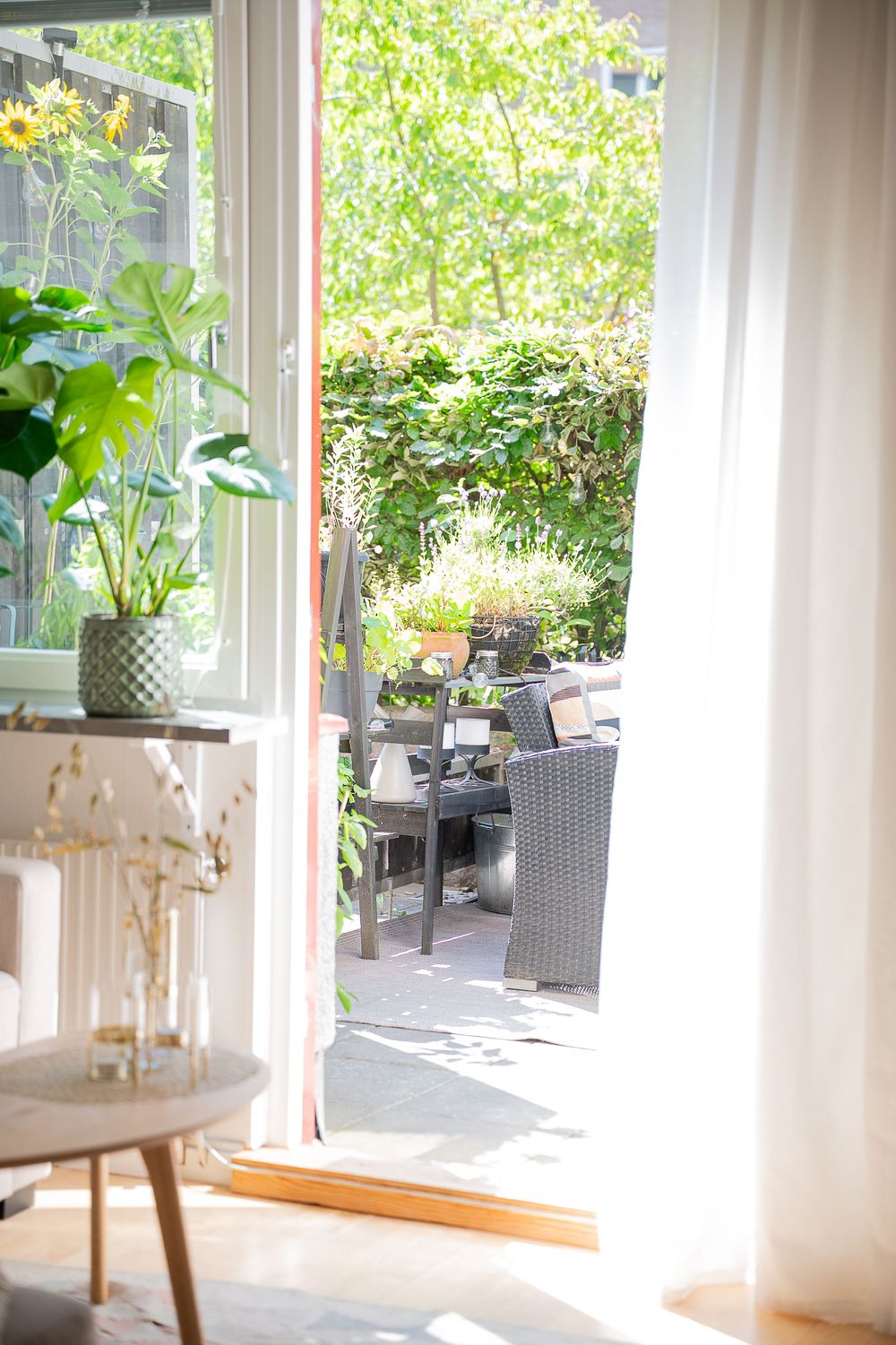 Open patio door reveals a cozy outdoor seating area with wicker furniture and lush greenery through white curtains.