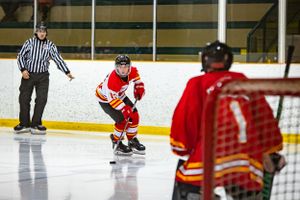 Hockey player in red jersey skates with puck while referee watches from side of rink.