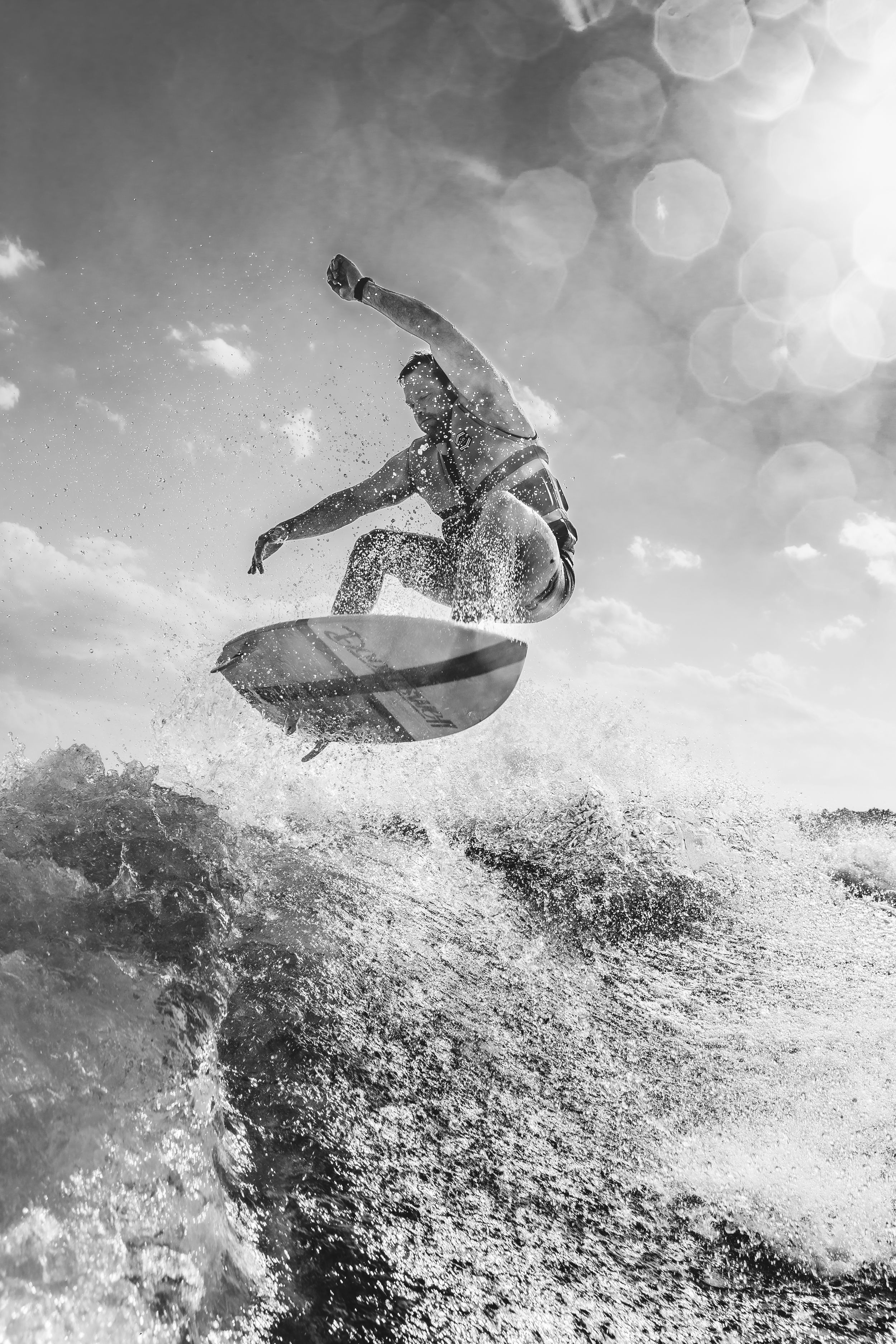 A surfer performs an aerial maneuver above the waves as water sprays in a dramatic black and white photo.