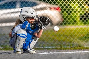 Young baseball catcher in blue uniform crouching behind home plate to catch incoming baseball.