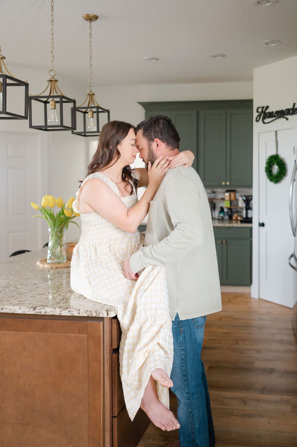 Intimate couple embracing in front of a sage green kitchen with pendant lighting.