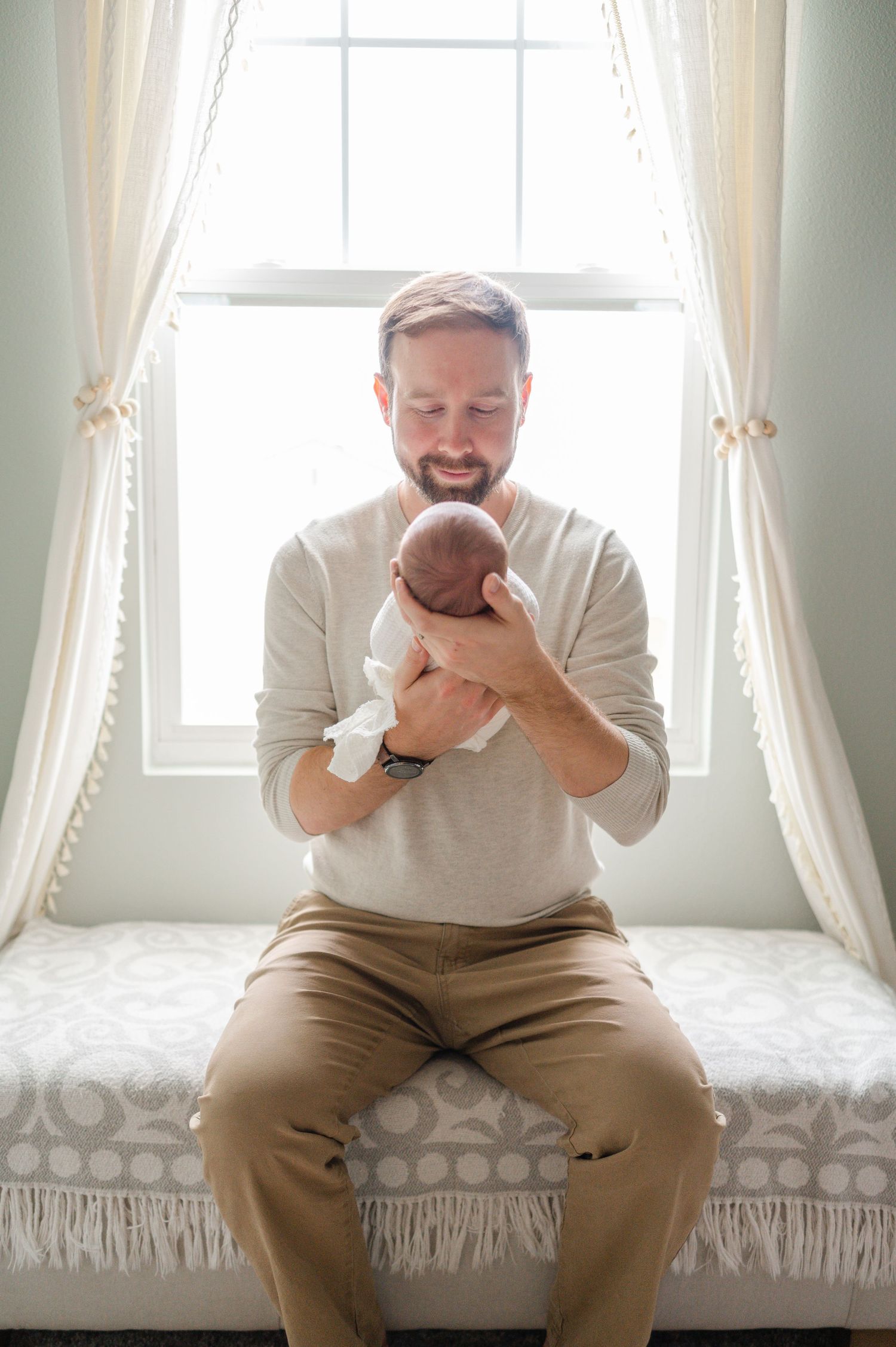 Parent sitting on window bench holding and smiling at their newborn baby.