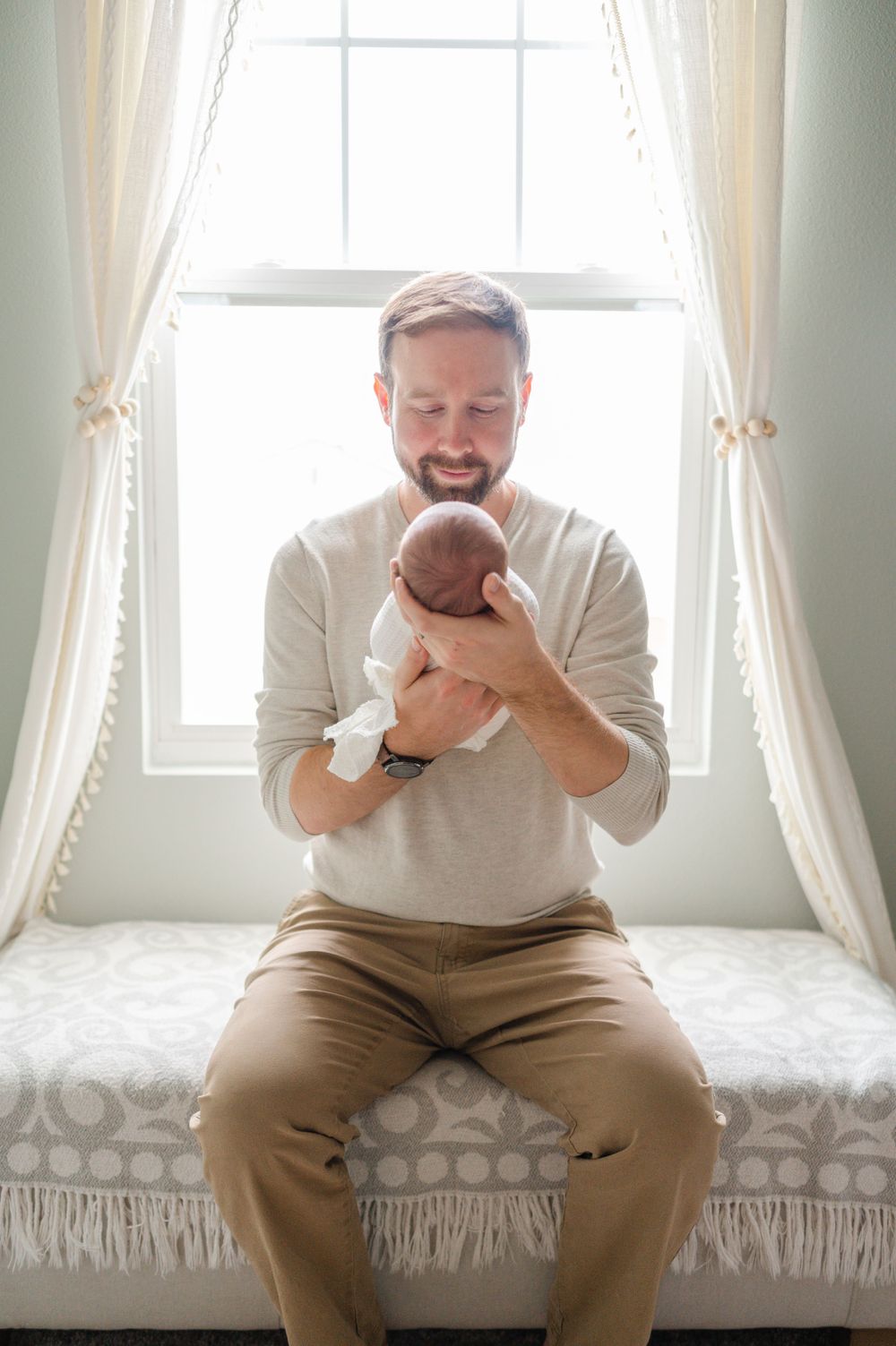Parent sitting on window bench holding and smiling at their newborn baby.