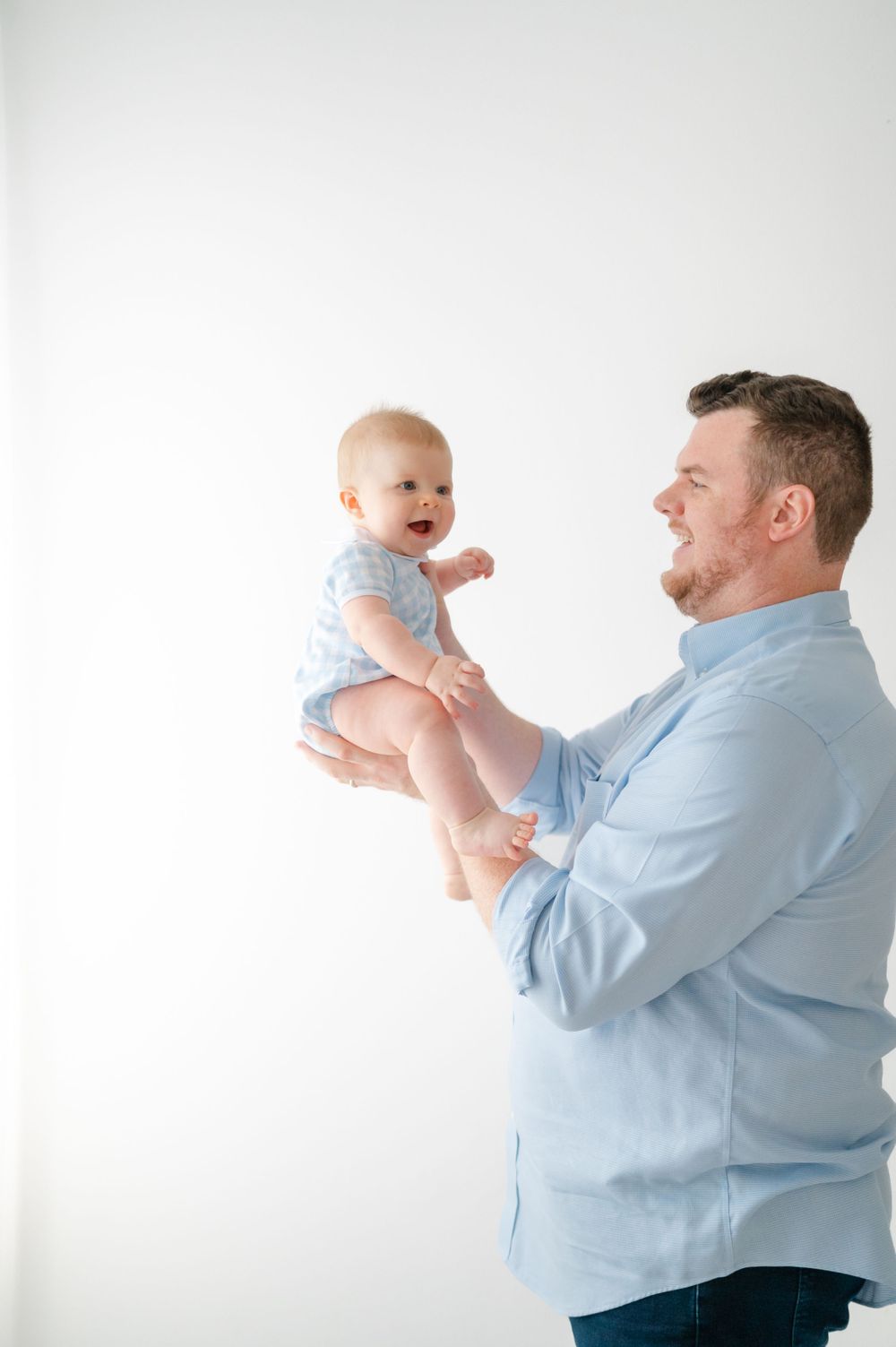 Baby in light blue outfit being lifted playfully against white background.