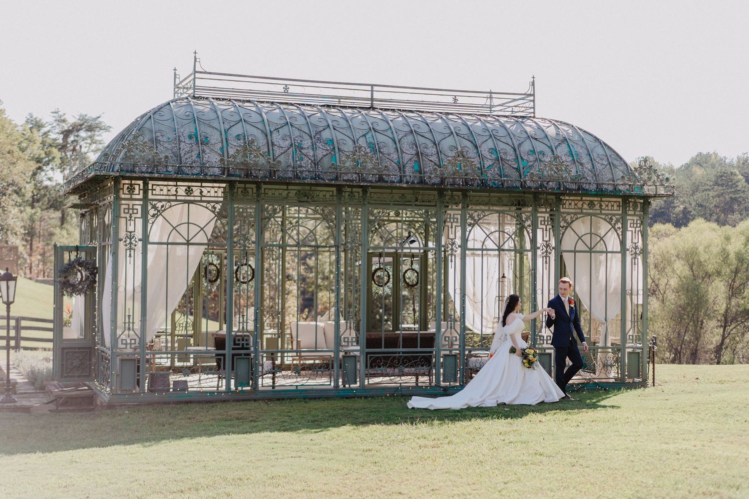 A romantic garden gazebo with ornate metalwork and glass panels sits on manicured lawn surrounded by trees.