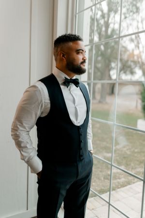 Elegant groom in formal attire with bow tie and waistcoat gazes thoughtfully through bright window on wedding day.