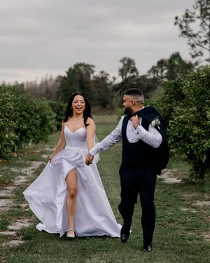 A couple in wedding attire walks and laughs together on a scenic outdoor path surrounded by greenery under overcast skies.