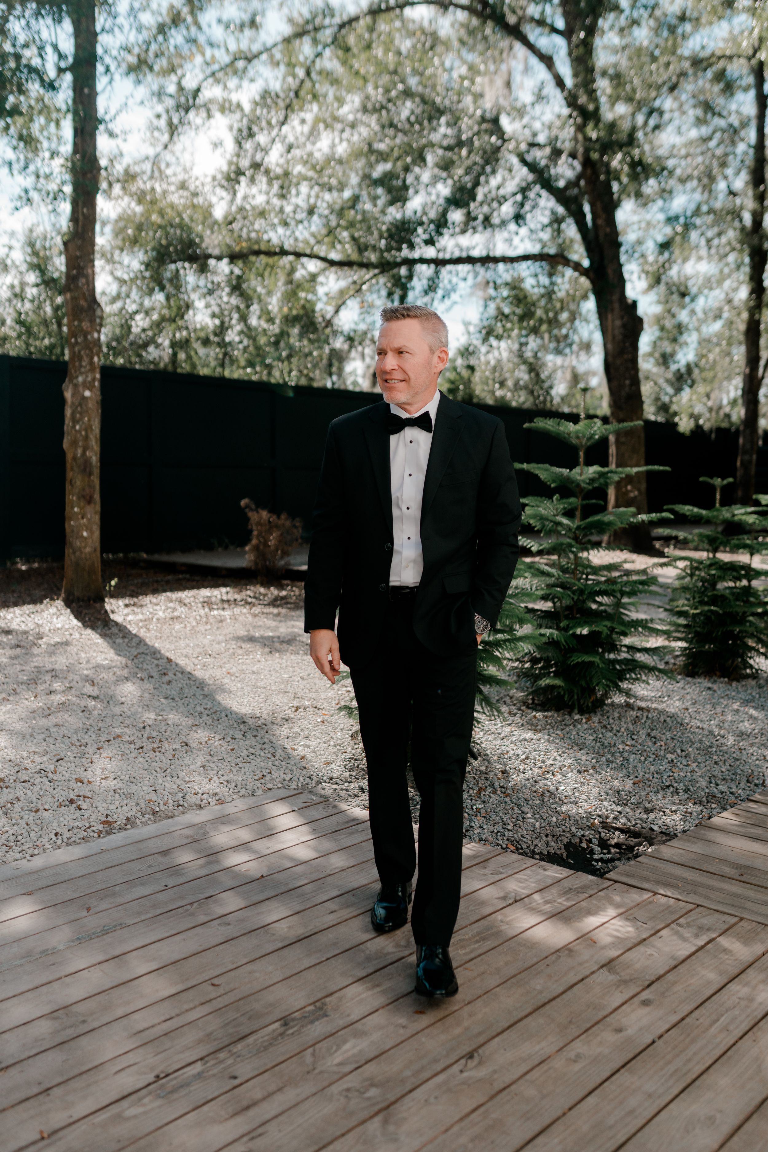 A person in a black suit walks across a wooden deck surrounded by trees in a natural outdoor setting.