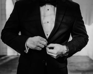 A black and white photo of someone adjusting their tuxedo bowtie in a formal setting.