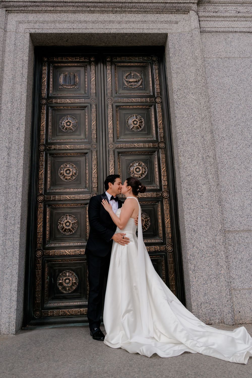Bride and groom kissing in front of ornate bronze doors in elegant wedding attire, white gown with train.