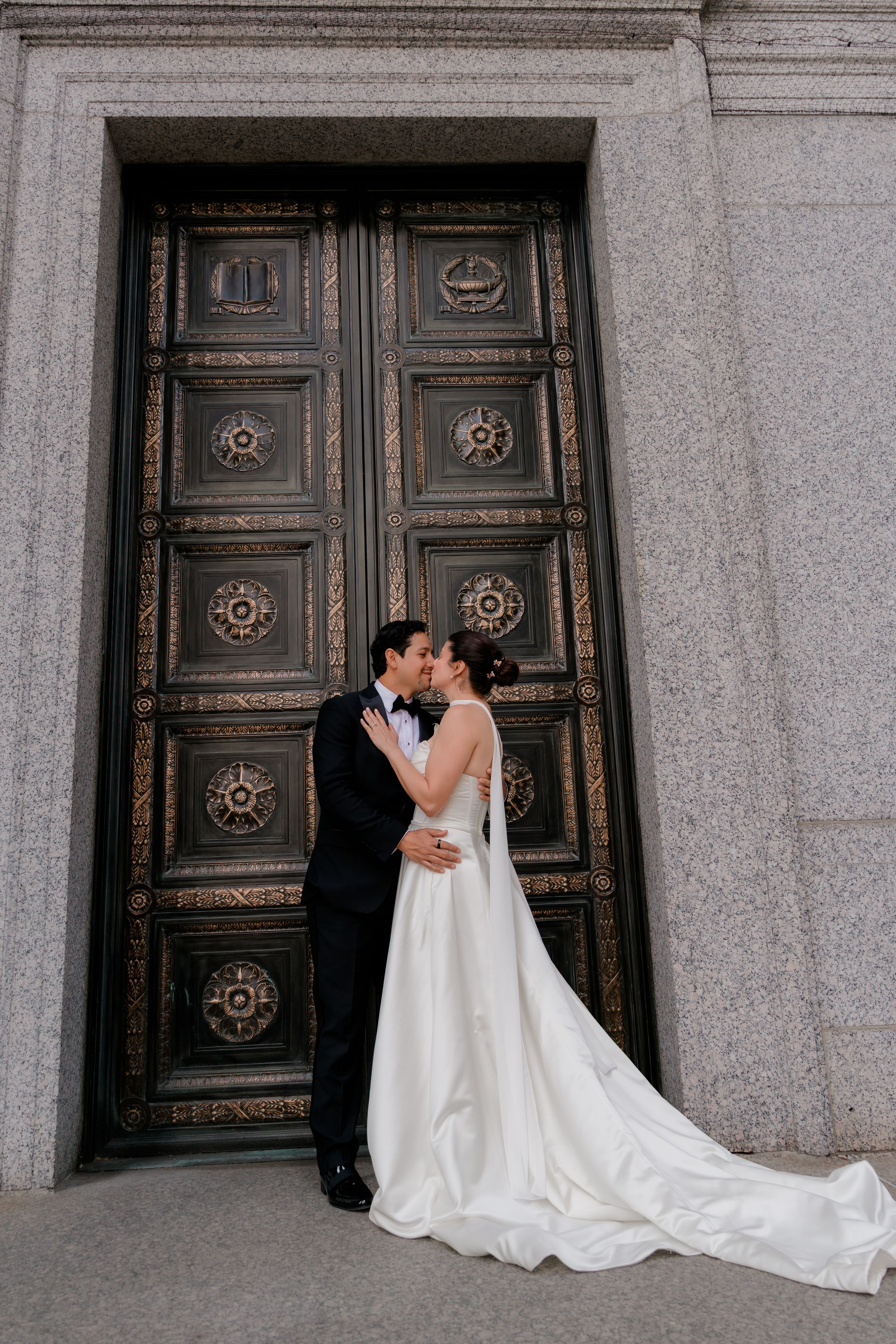 Bride and groom kissing in front of ornate bronze doors in elegant wedding attire, white gown with train.