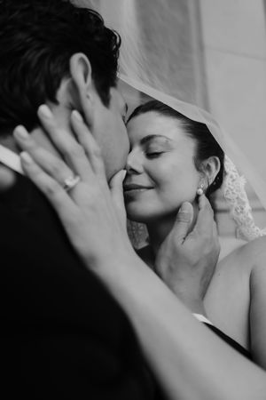 Black and white wedding photo of groom tenderly holding bride's face for a kiss, bride wearing veil and pearl earrings.