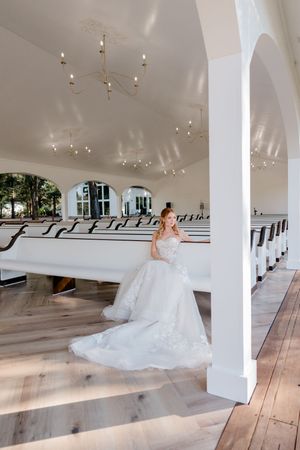Bride in a flowing white lace gown poses beside a white column in an elegant chapel with wooden pews.