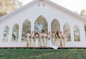 A wedding party stands in front of a white chapel with Gothic arched windows in a natural outdoor setting.