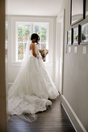 A flowing white wedding dress creates a dreamy silhouette in a bright hallway with natural light streaming through windows.