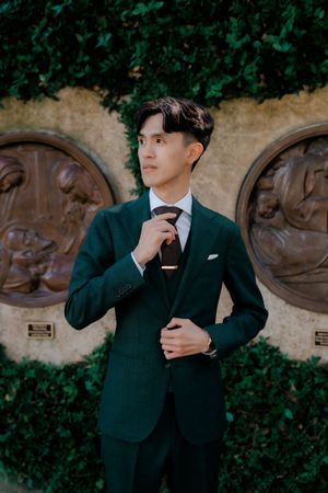 A person in a dark suit adjusts their tie and cuffs while posing against a wall with climbing vines and stone carvings.