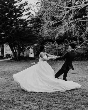 A bride and groom dance playfully outdoors as her white wedding dress twirls dramatically in black and white photography.