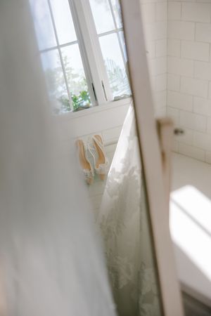 A pair of elegant beige high heel shoes sit by a bright window in a white bathroom with natural sunlight streaming in.