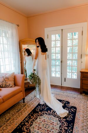 Elegant white wedding dress with long train shown in sunlit room with French doors and vintage furniture on patterned rug.