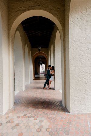 Long arched corridor with white stone walls and terracotta hexagonal tile flooring leads to distant figures in window alcoves.