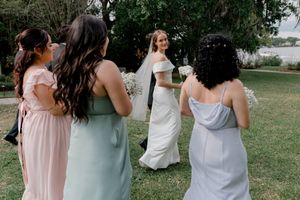 A bride in a white wedding gown walks on grass while bridesmaids in pastel dresses look on during an outdoor celebration.