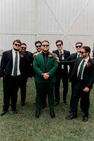 A groom in emerald green suit stands with groomsmen wearing black suits and sunglasses for wedding party photo.