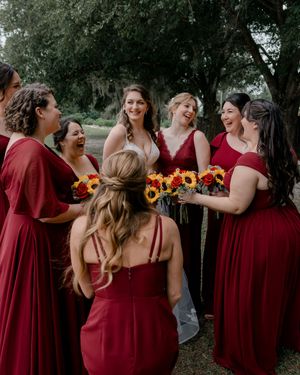 Bridesmaids in burgundy dresses gather outdoors holding vibrant sunflower bouquets at a wedding celebration.