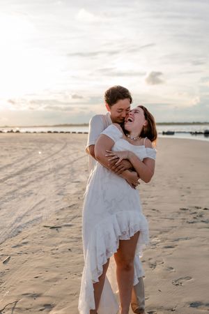 A romantic couple in white outfits share tender moments together on a sandy beach at sunset.