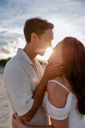 A romantic couple embraces on the beach at sunset with soft sunlight glowing behind them.