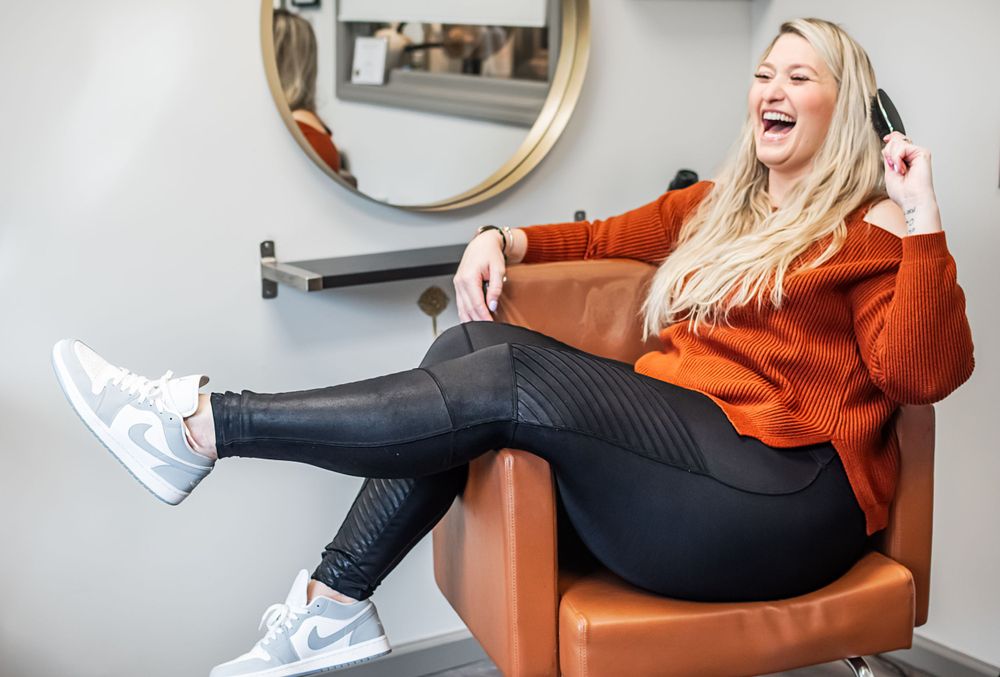 Woman in orange sweater laughs while sitting in leather chair against white wall with mirror.