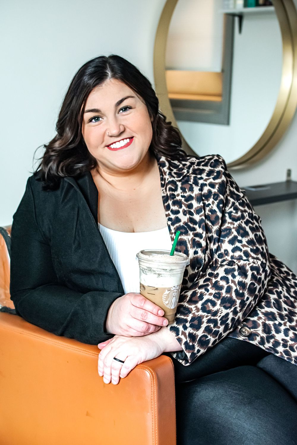 Woman in leopard print blazer holding coffee drink while sitting on orange chair near circular mirror.