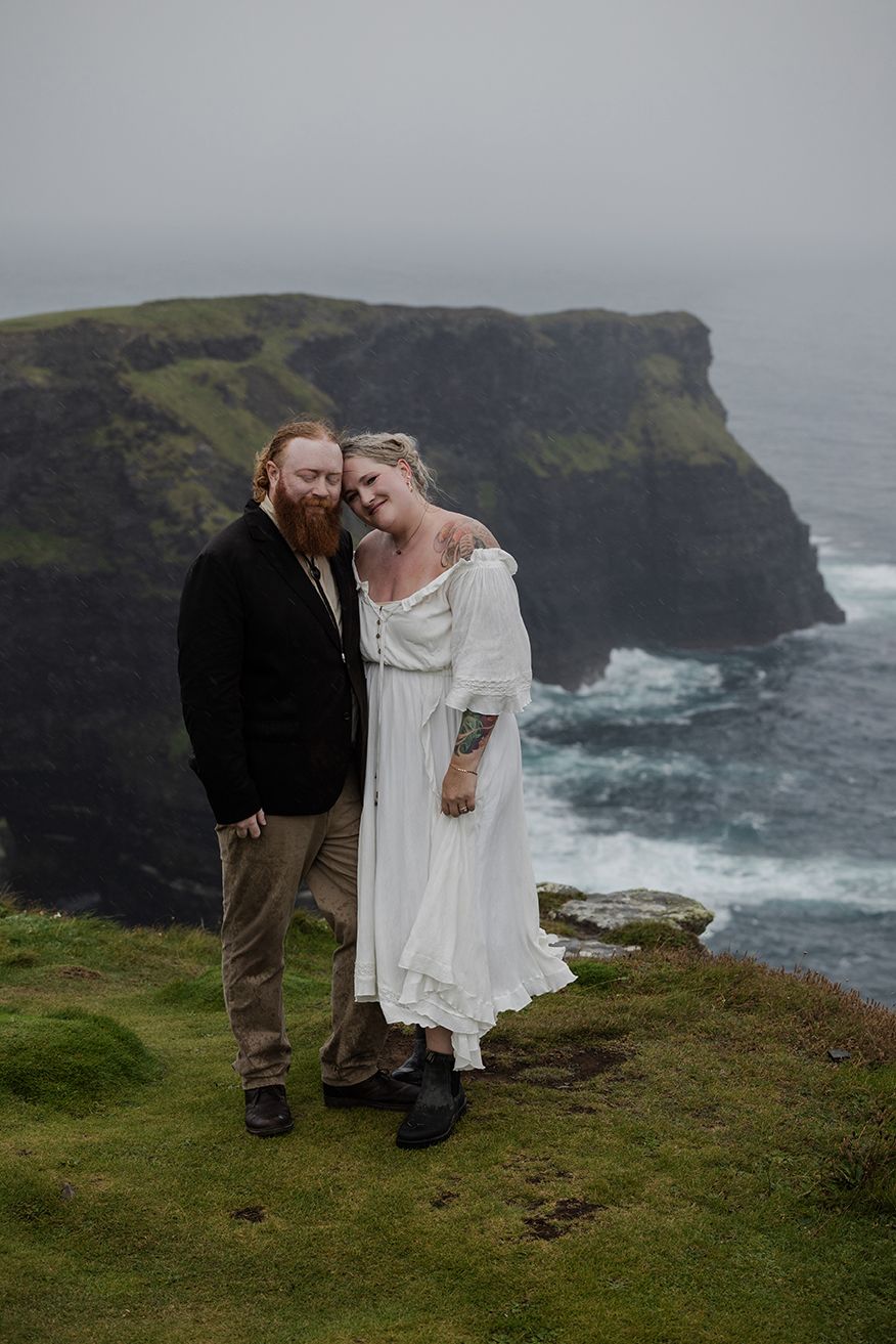 Couple standing, holding each other close, by the Cliffs of Moher during their adventure elopement in Ireland