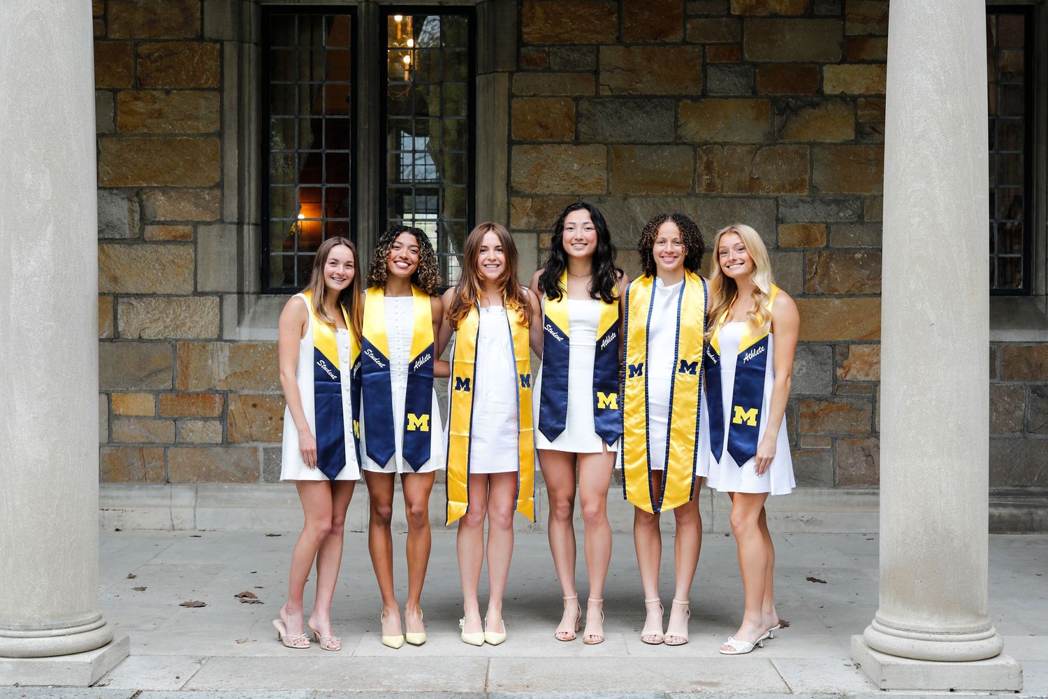 Six female graduates in University of Michigan regalia posing together at stone campus building.