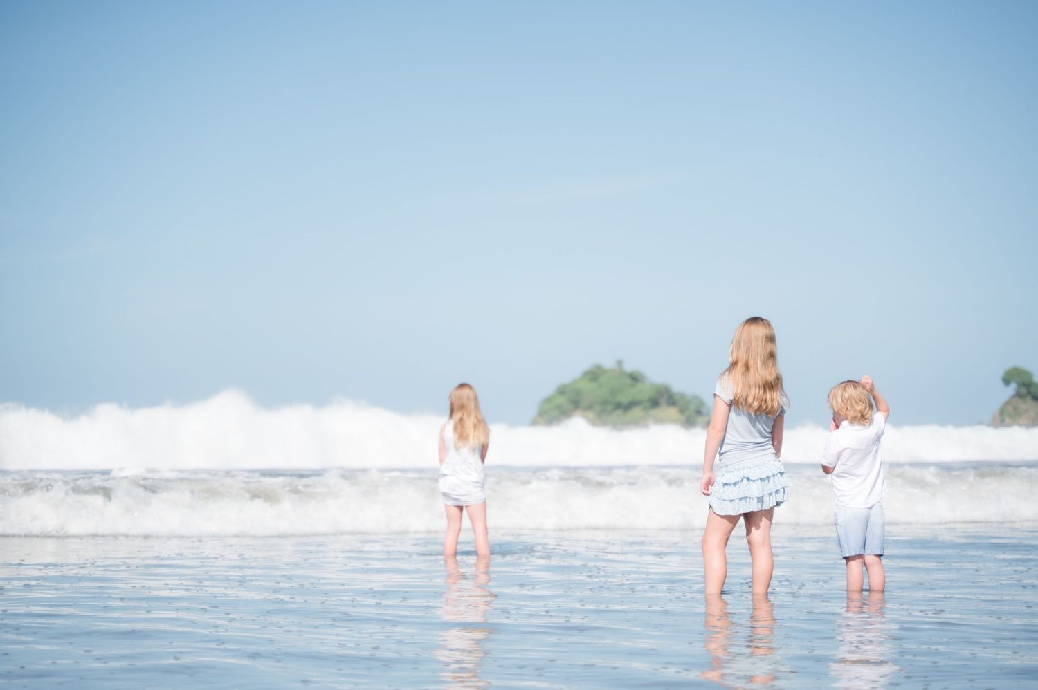 Three children standing at the shoreline of a tropical beach, facing ocean waves with reflections on wet sand.
