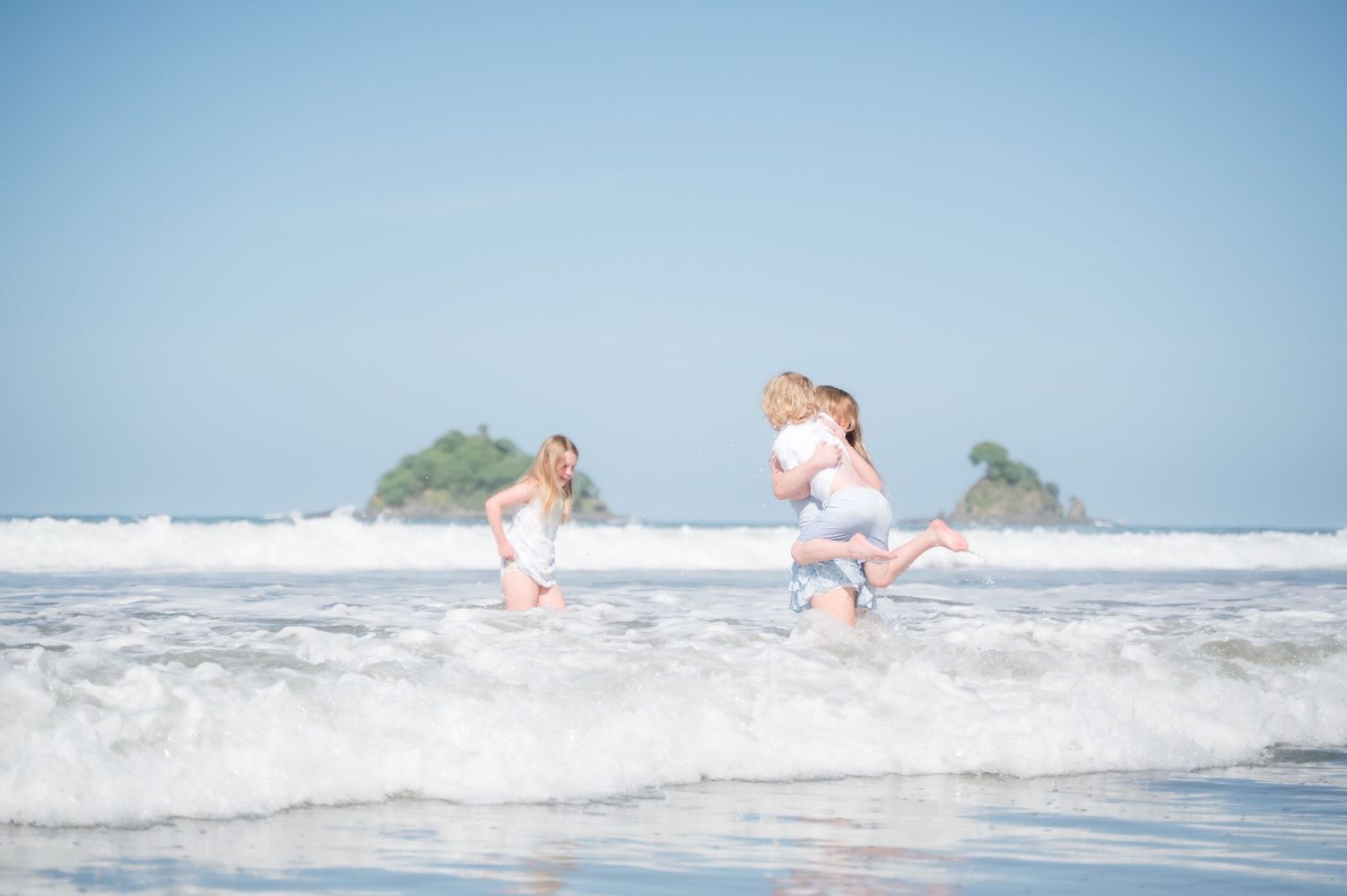 Three children playing joyfully in ocean waves on a tropical beach with small islands visible in the background.