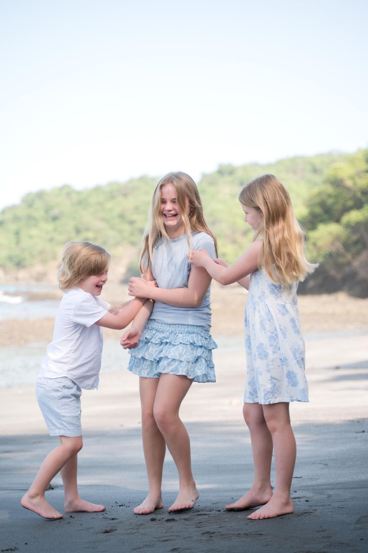 Three barefoot children laughing and playing together on a sandy beach with green hills in the background.
