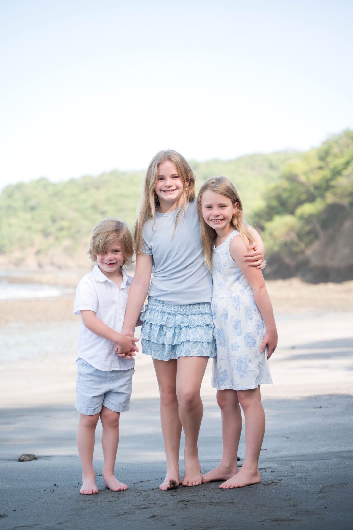 Three barefoot children pose together on a sandy beach, smiling in light blue and white outfits.