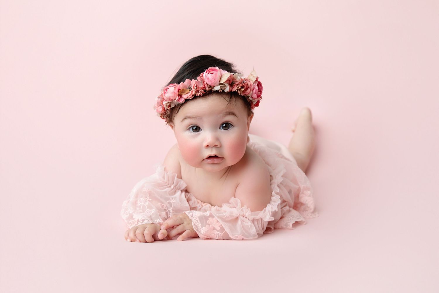 Baby in mint green dress seated in vintage wicker chair against neutral background.