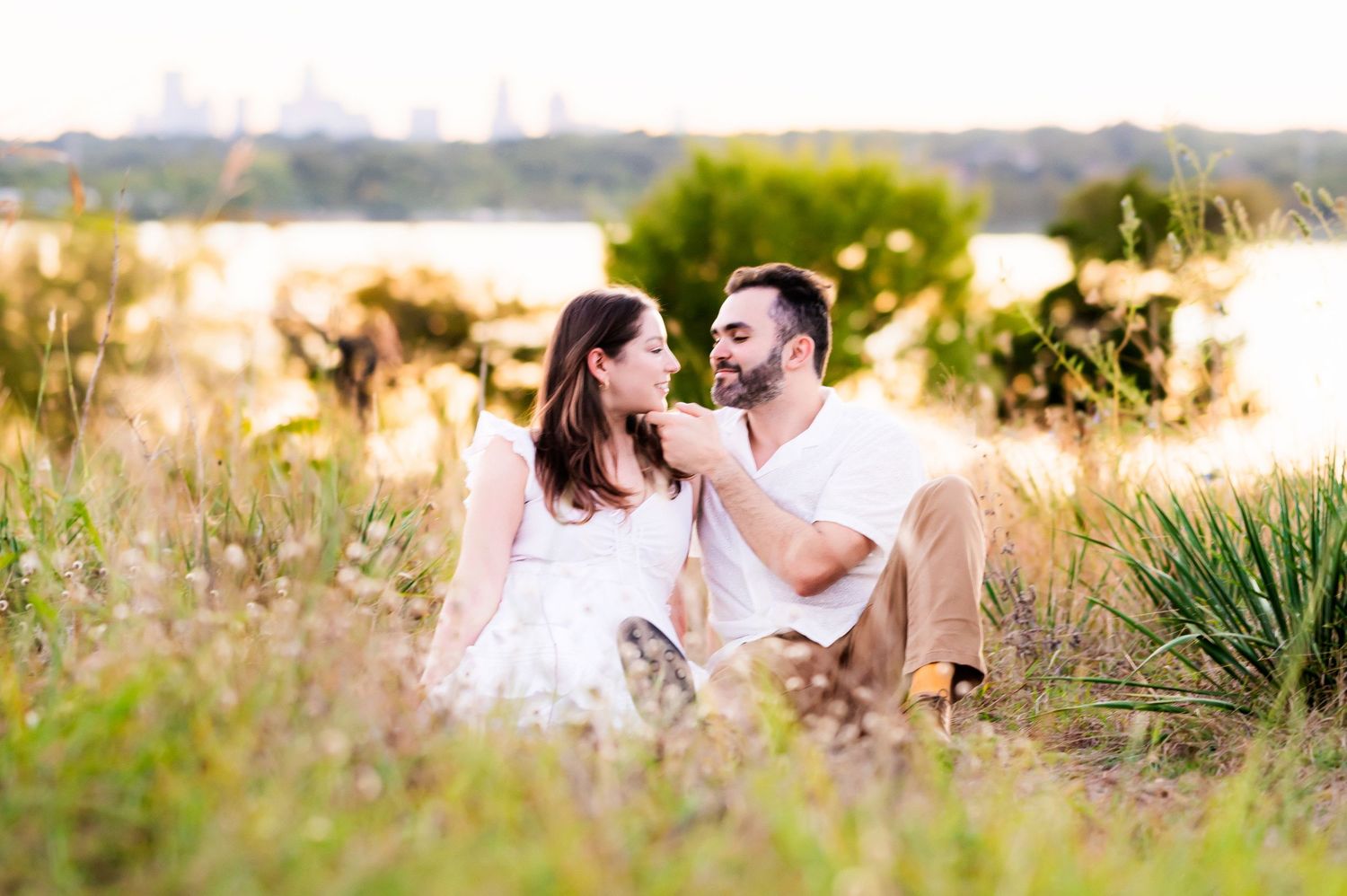 East Dallas couple posing seated among wild grasses for engagement photos at Winfrey Point, white rock lake and dallas skyline in background