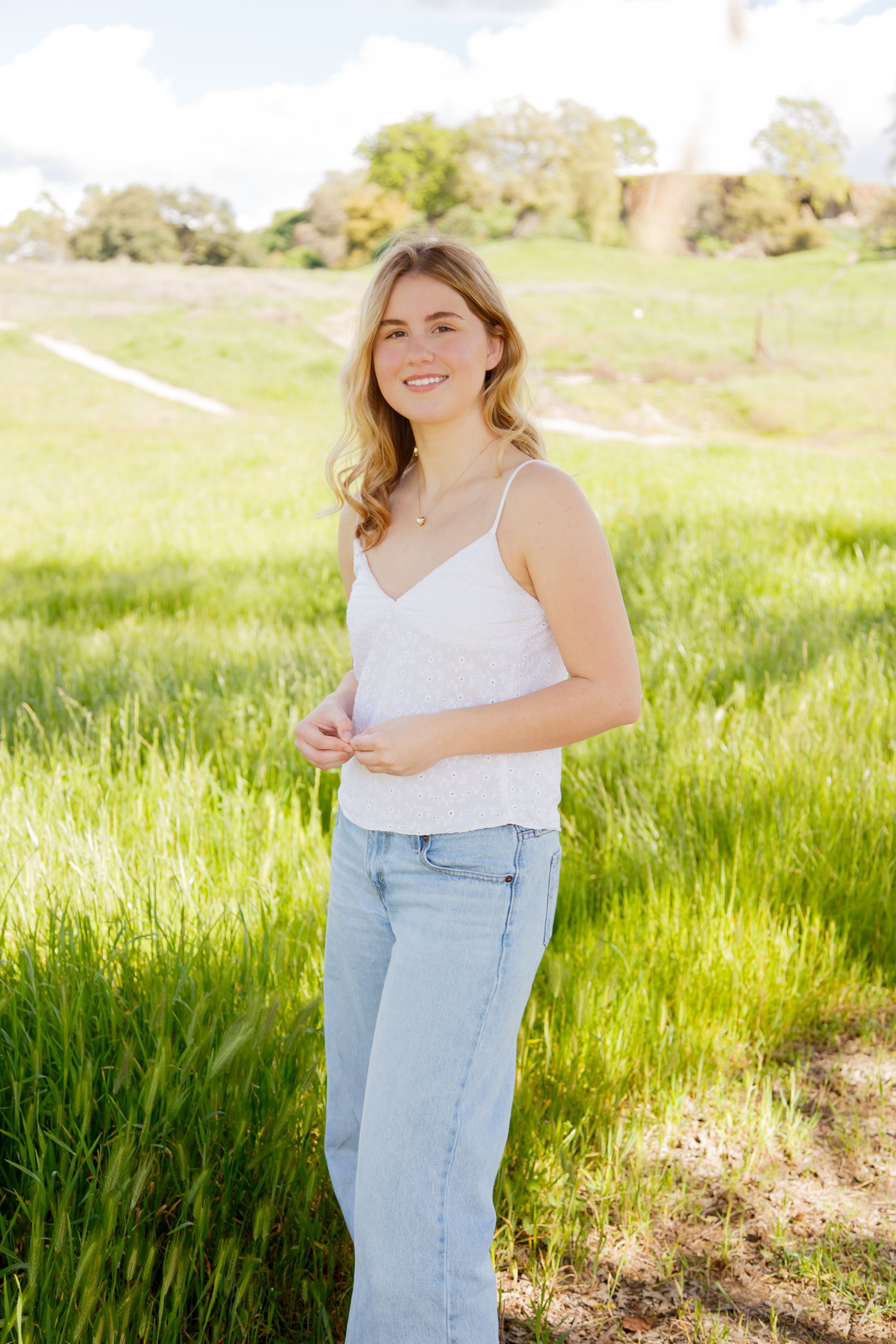 Young blonde woman smiling in white tank top and jeans, standing in a sunny green field.