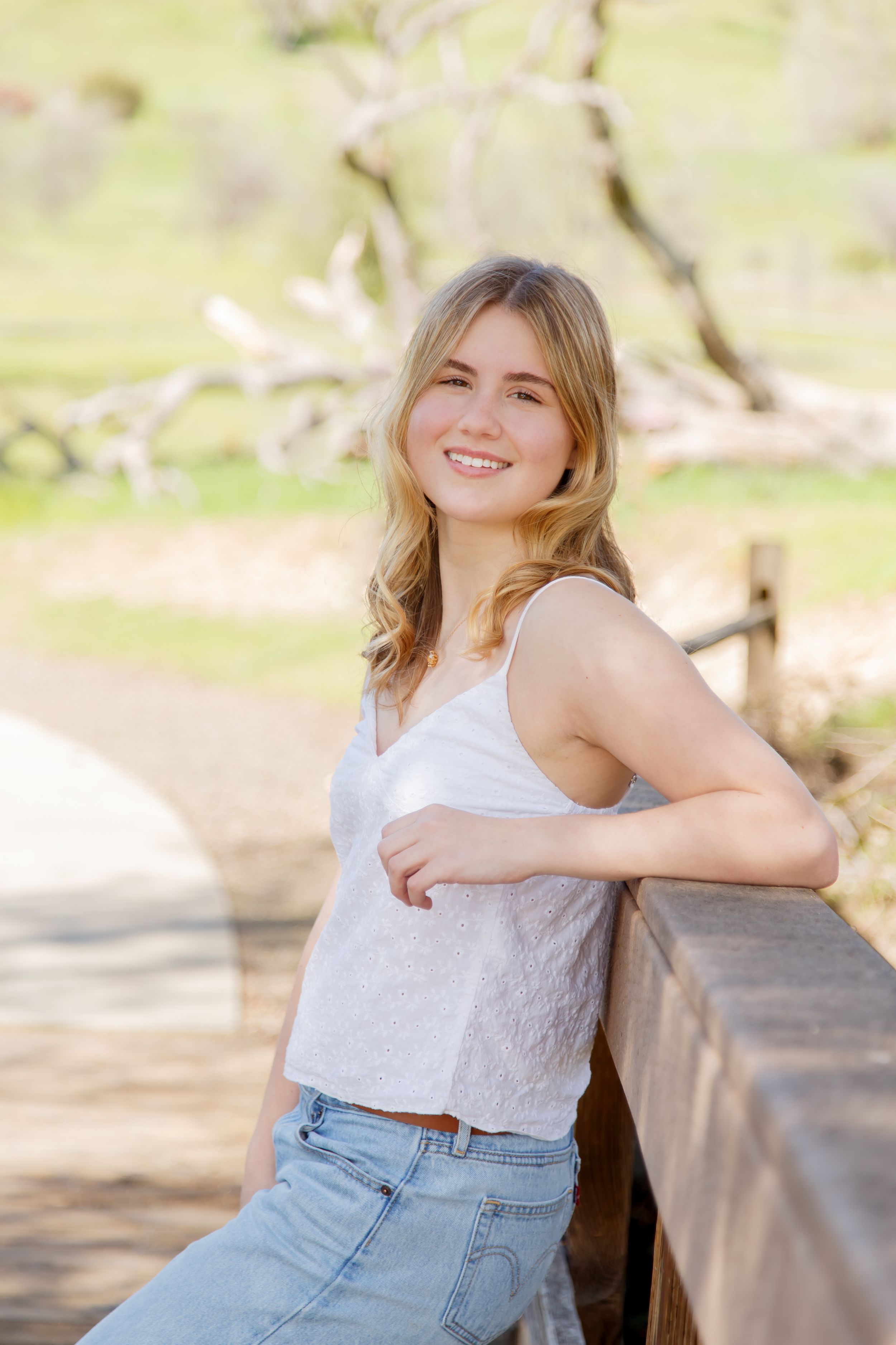 Smiling young blonde woman in white tank top and jeans leaning on wooden fence in sunny outdoor park.