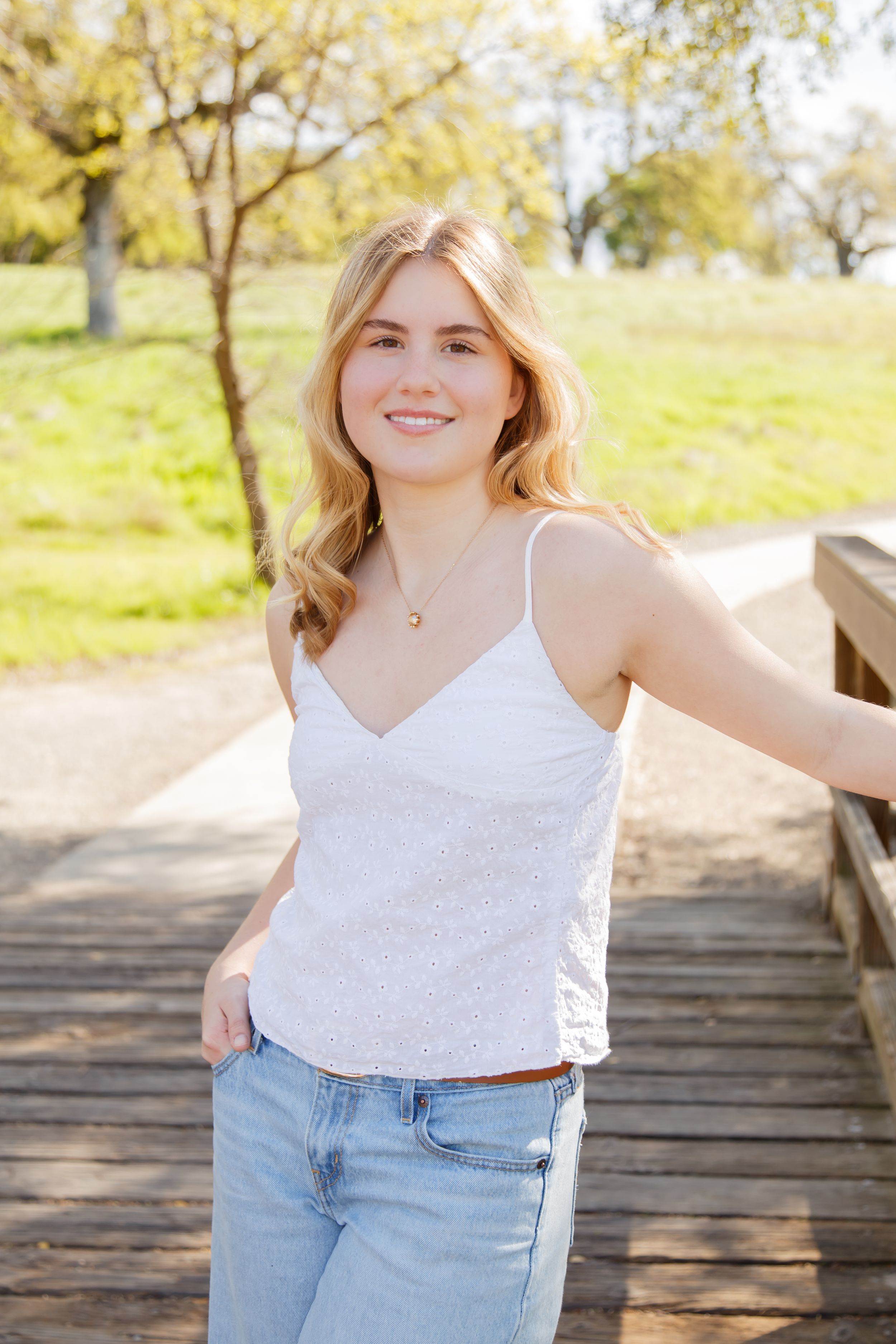 Young blonde woman in white tank top and jeans smiles on a wooden bridge in a sunny park.