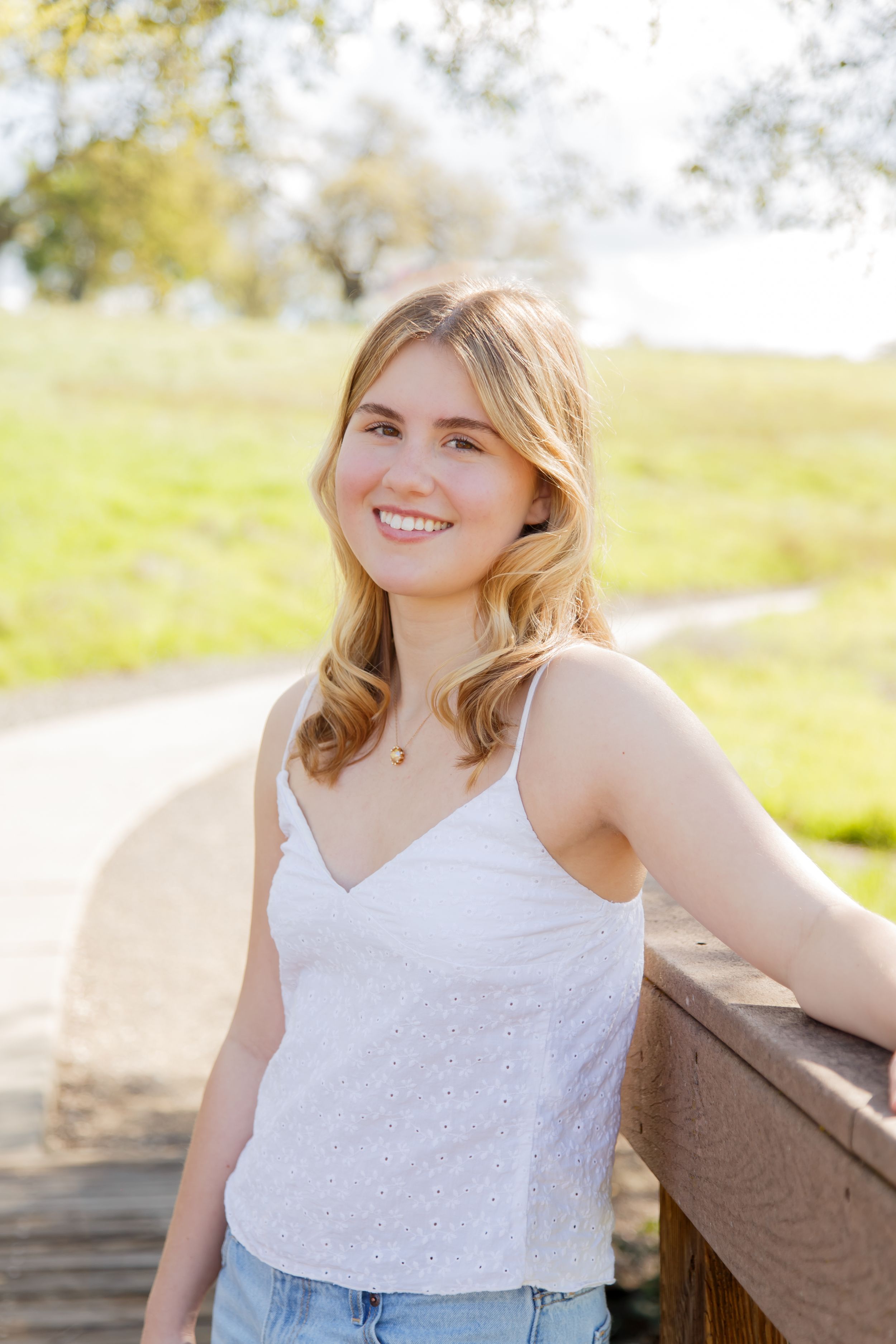 Smiling young blonde woman in white eyelet tank top leaning on wooden railing in sunny park setting.